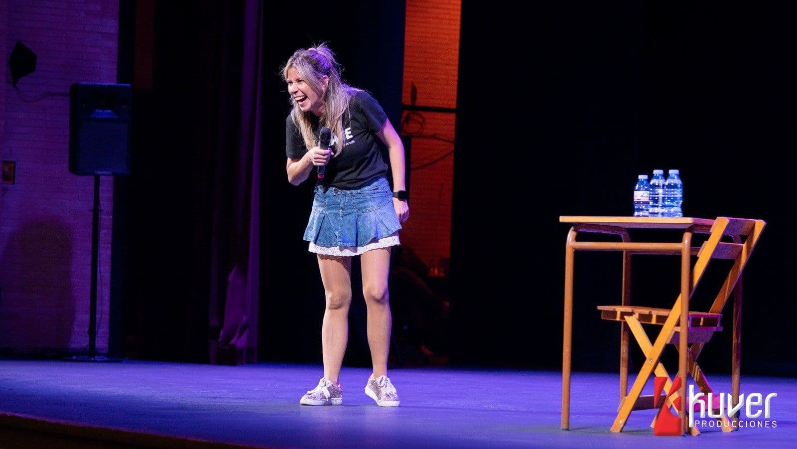 Palo Capilla haciendo stand up en un escenario de teatro con una mesa y botellas de agua