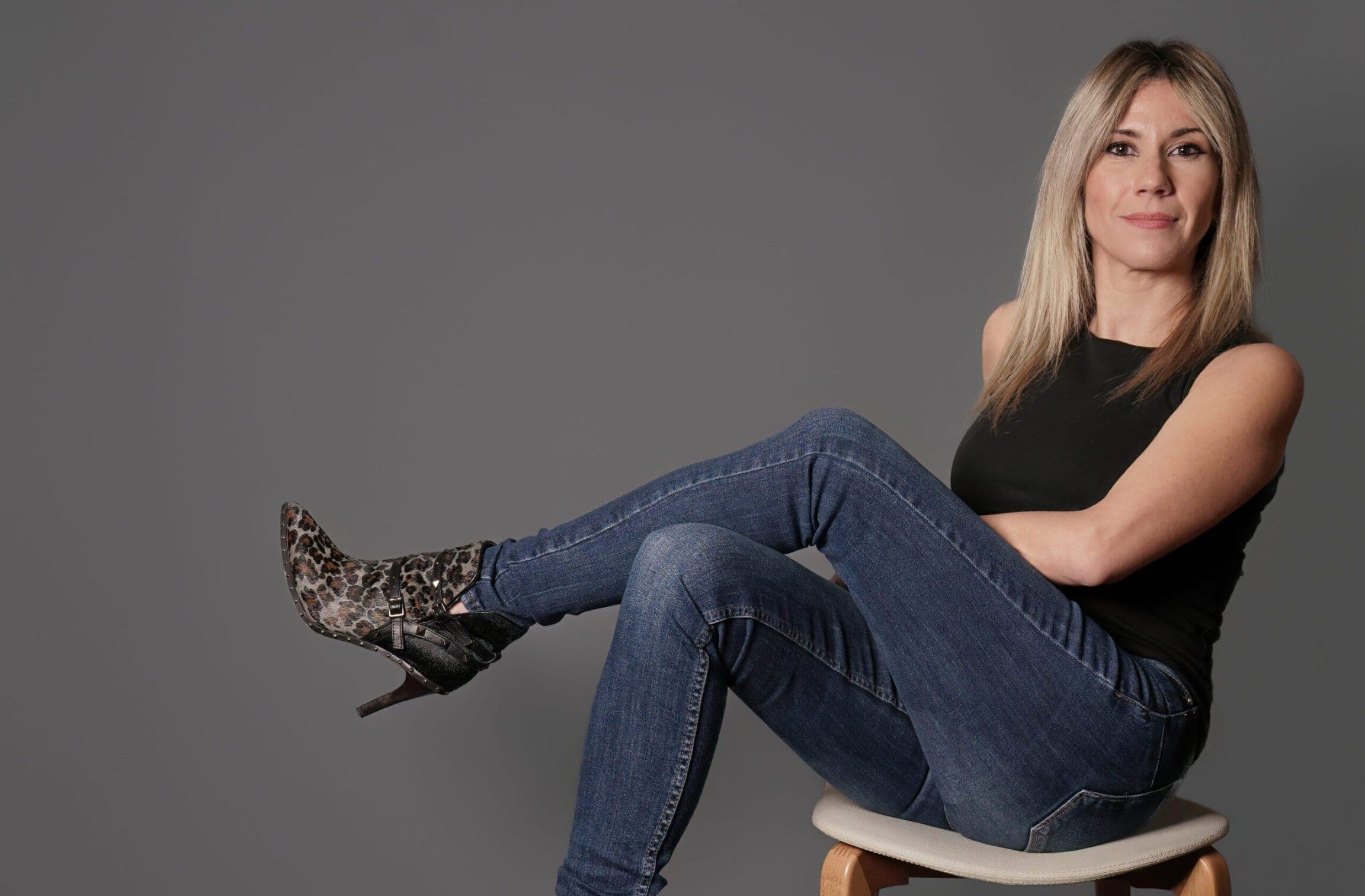 Paloma Capilla sentada en un taburete con vaqueros y botas de leopardo, fondo gris